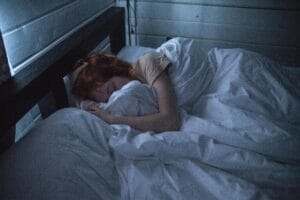 7-Day Reset Plan 6 | eat move healthy A woman sleeping peacefully in a cozy bedroom, enveloped by soft white sheets, under the gentle glow of night lighting.